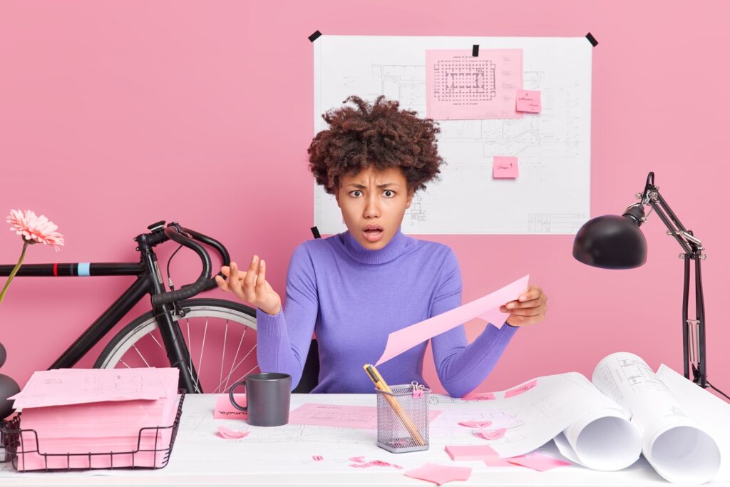 irritated puzzled ethnic female engineer holds paper raises hand has displeased face expression works in office prepares project work isolated over pink background. people and occupation concept