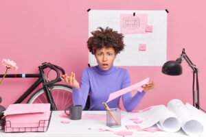 irritated puzzled ethnic female engineer holds paper raises hand has displeased face expression works in office prepares project work isolated over pink background. people and occupation concept